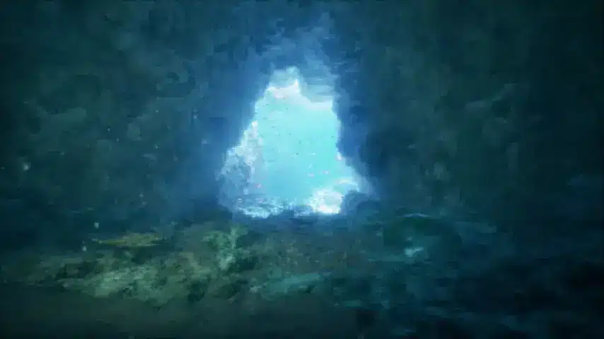 - N9BO℠ | Global Underwater Services Ltd Underwater photo showing the view from inside a rocky cave or tunnel, looking out towards bright blue water illuminated by sunlight, with bubbles and rocks visible around the entrance.