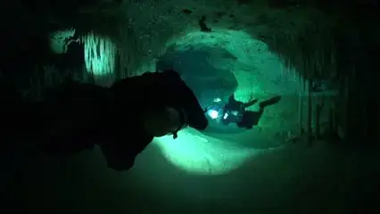 Two scuba divers with torches explore an underwater cave, surrounded by rock formations and illuminated by their lights in the dark, green-tinted water.