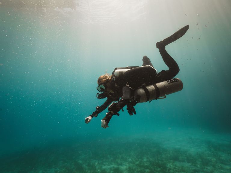 A scuba diver wearing diving kit and an oxygen cylinder swims underwater, surrounded by clear blue-green water and faint sunlight streaming from above.