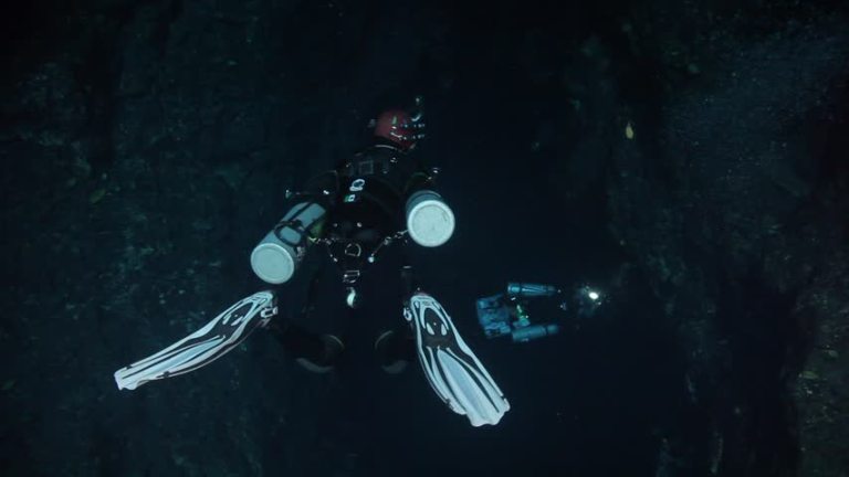 Two scuba divers wearing wetsuits, fins, helmets, and carrying cylinders swim in dark underwater surroundings, illuminated by a diver’s torch.