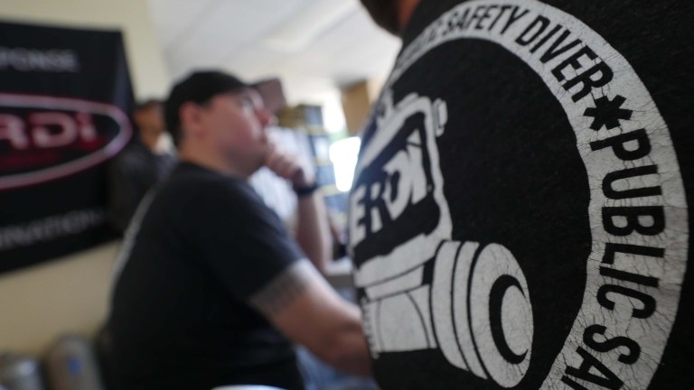 A close-up of a person wearing a shirt with a PUBLIC SAFETY DIVER logo; in the background, another person sits at a table, partially out of focus, appearing to listen or think.