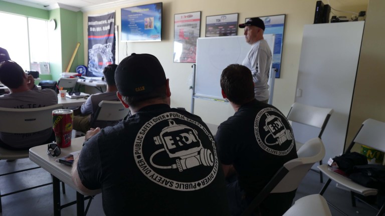 A group of people sit in a classroom facing a man standing at a whiteboard. Several attendees wear black shirts with a circular “ERDI Public Safety Diver” logo on the back. Posters hang on the walls.