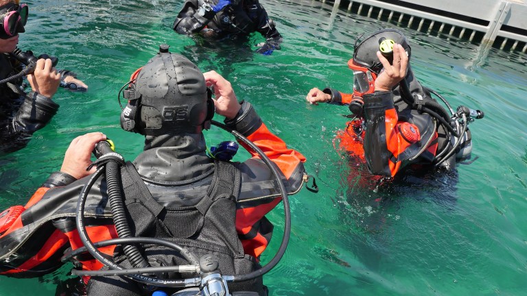 Four scuba divers in wetsuits and full kit are preparing in clear, turquoise water near a metal jetty. One diver faces the camera, adjusting equipment, while others interact and adjust their masks.