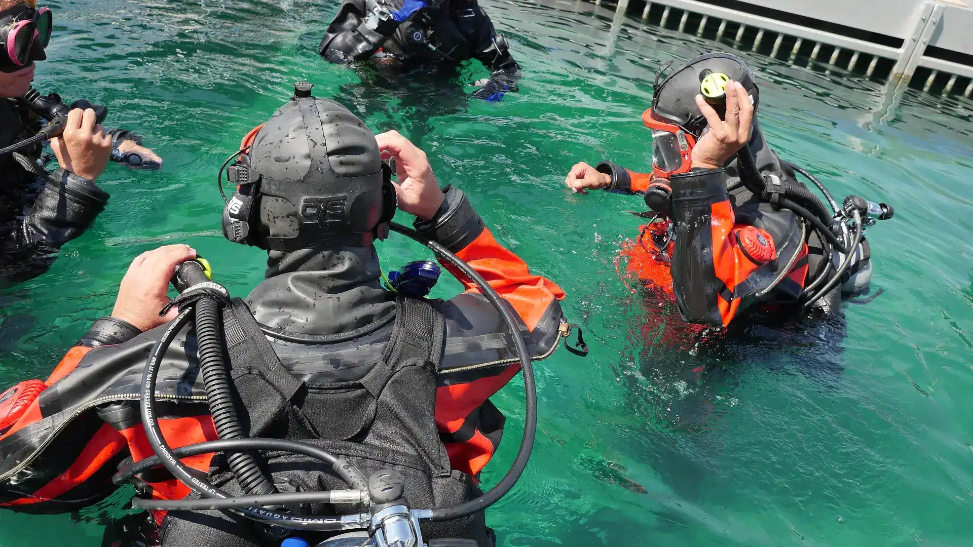 Four scuba divers in wetsuits and full kit are preparing in clear, turquoise water near a metal jetty. One diver faces the camera, adjusting equipment, while others interact and adjust their masks.
