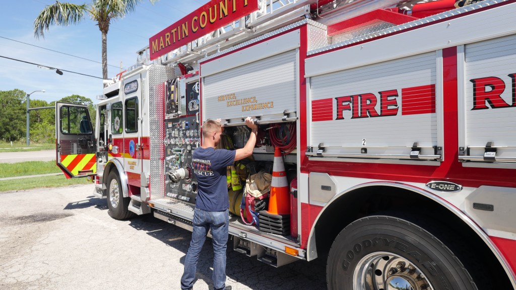 ERDI-Fire-Truck-Photo - N9BO℠ | Global Underwater Services Ltd A firefighter in a navy shirt stands beside a Martin County Fire and Rescue lorry, organising equipment such as hoses and orange cones stored in a side compartment.