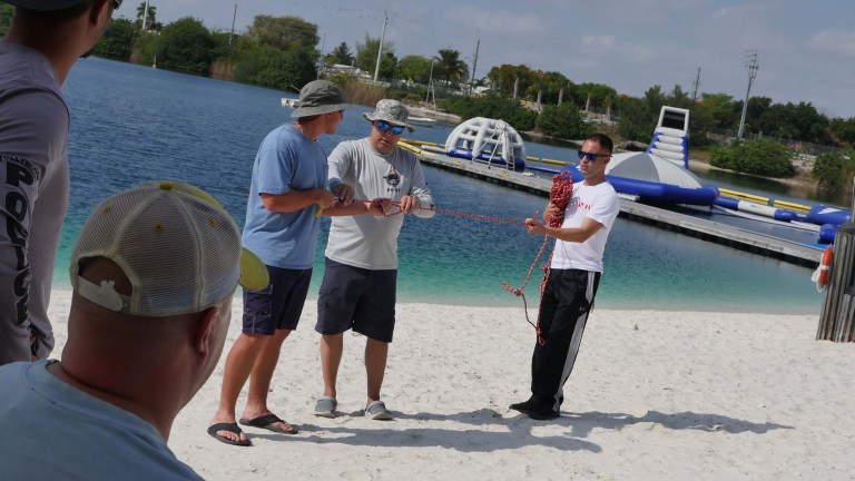 Three men stand on a sandy beach near a lake with inflatable water structures. Two men hold a rope, whilst the man on the right holds a coiled section. Other people watch from the foreground.