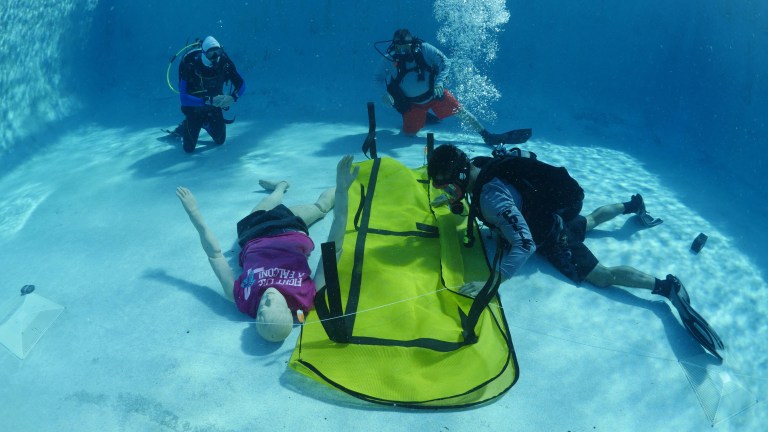 Four scuba divers practise an underwater rescue drill in a swimming pool, positioning a yellow stretcher next to a mannequin lying face-up, whilst one diver photographs the scene.
