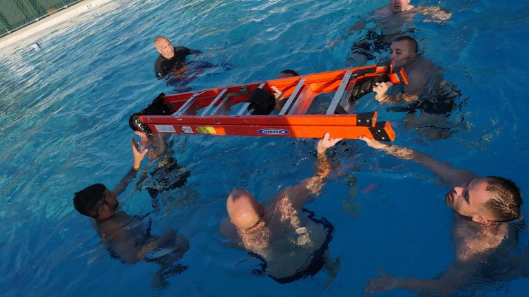 Several people in a swimming pool hold and balance an orange ladder horizontally above the water, working together as a group. The setting appears to be a swimming or rescue training exercise.