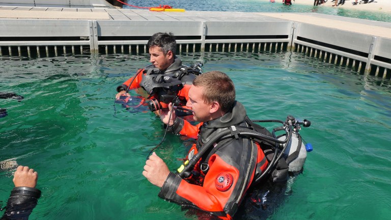 Two scuba divers in orange dry suits are standing in clear green water near a dock, preparing their kit. One holds a mask while both wear oxygen cylinders and other diving equipment.