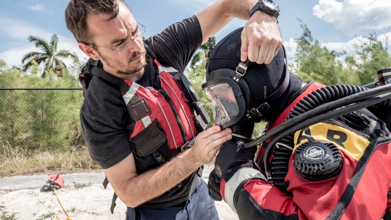 A man in a red life jacket helps a diver in full scuba gear adjust their face mask outdoors, with trees and a blue sky in the background.
