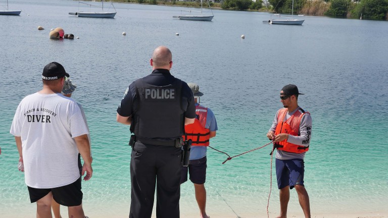 Four men stand by the shore of a lake; one wears a police vest, one a shirt labelled diver, and two others in life vests handle a rope. Sailing boats are visible on the water in the background.