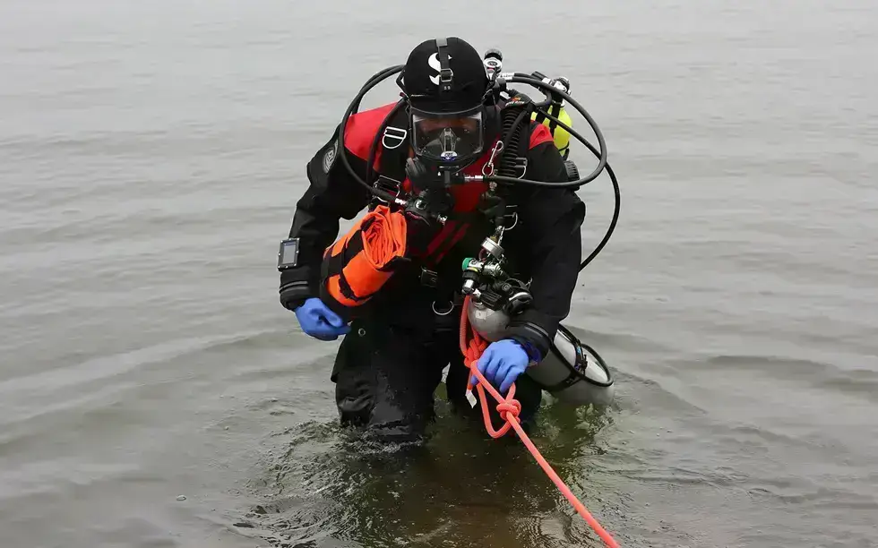 A scuba diver wearing a black and red wetsuit, blue gloves, and full kit emerges from the water holding an orange flotation device attached to a rope.