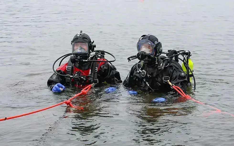 Two scuba divers in full kit and drysuits stand waist-deep in the water, each holding onto red safety ropes, with their masks and equipment clearly visible.