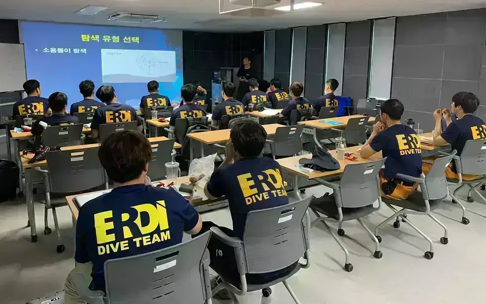 A classroom with several people wearing “ERDI DIVE TEAM” shirts sit at desks facing a screen. An instructor stands at the front, presenting slides with Korean text and diagrams.
