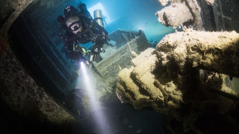 A scuba diver in full kit shines a torch while exploring the interior of a sunken shipwreck underwater, surrounded by corroded metal and marine growth.