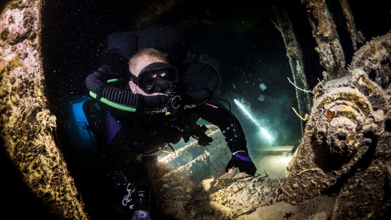 A scuba diver in full kit explores the interior of a sunken shipwreck underwater, shining a torch and surrounded by rusted metal and debris in a dark, mysterious environment.