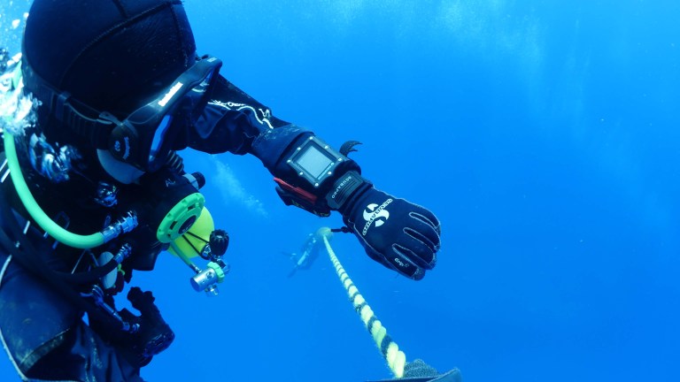 A scuba diver in a wetsuit and gloves holds onto a yellow rope underwater, whilst wearing a dive computer on their wrist. The deep blue ocean surrounds them, with another diver visible in the distance.