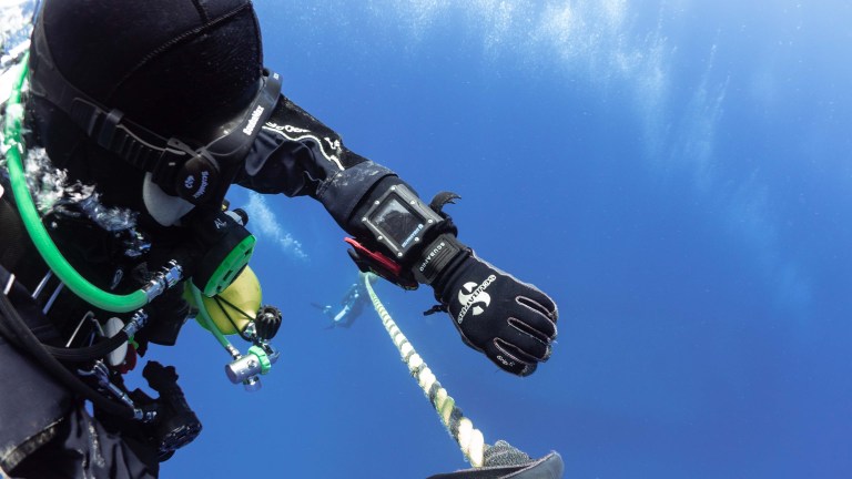 A scuba diver in full kit holds onto a rope while underwater, with blue water surrounding them. The diver wears a dive computer on their wrist and has a green cylinder and camera attached to their suit.