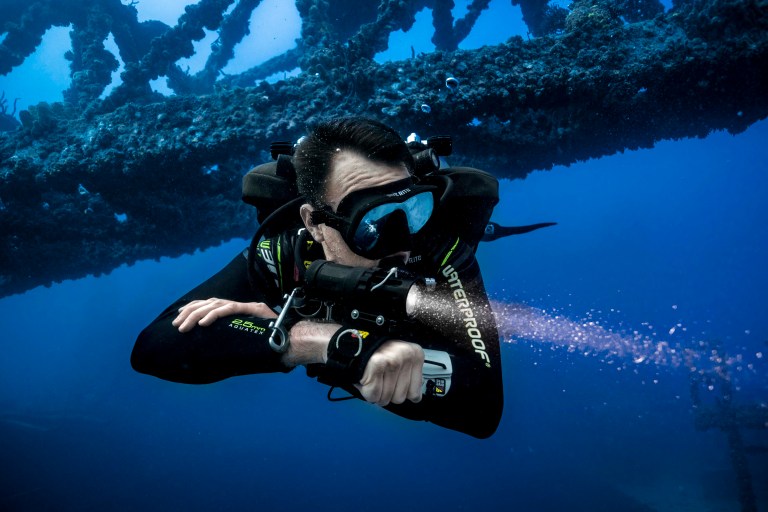 A scuba diver in a black wetsuit and goggles swims underwater near a large, encrusted shipwreck structure, releasing a stream of bubbles whilst surrounded by deep blue water.