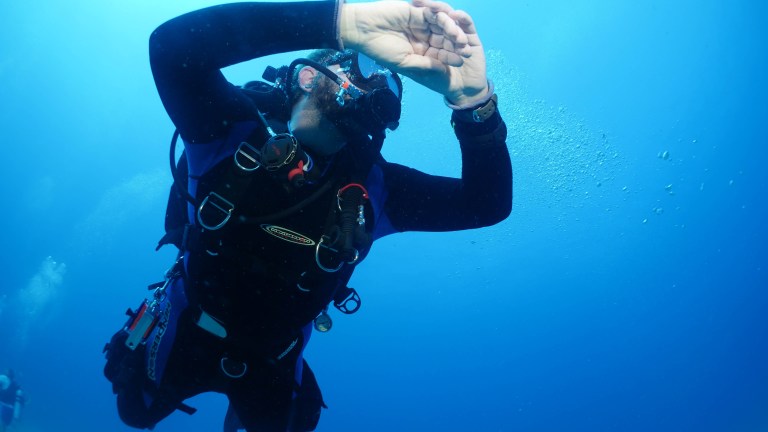 A scuba diver underwater wearing a black wetsuit and diving kit, holding their hand in front of their face whilst looking up, surrounded by clear blue water and bubbles.