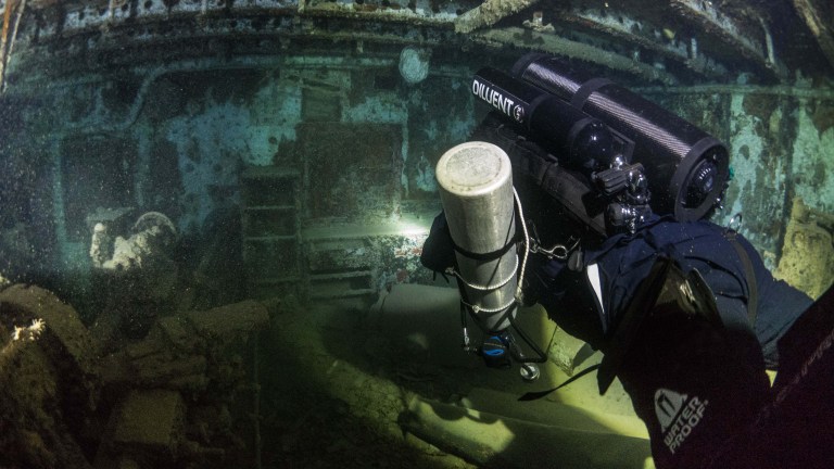 A scuba diver with advanced gear explores the interior of a sunken shipwreck underwater, shining a torch to illuminate debris and corroded structures in a murky environment.