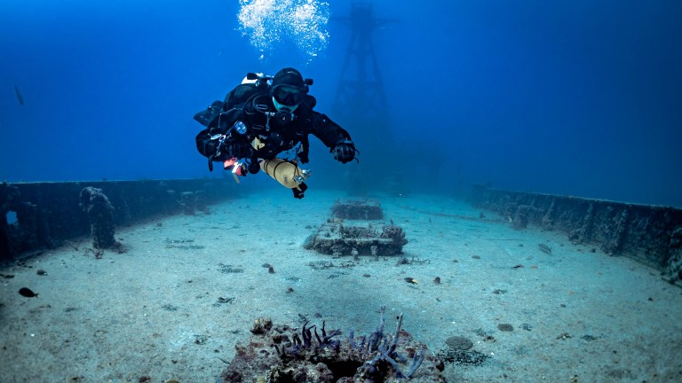 A scuba diver swims above the sandy floor of a sunken shipwreck under clear blue water, with bubbles rising and a large underwater structure visible in the background.