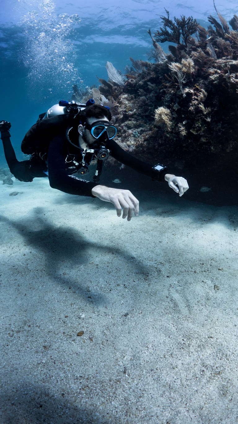 A scuba diver in full kit swims near the sandy sea floor beside a coral reef, with sunlight filtering through the water above.
