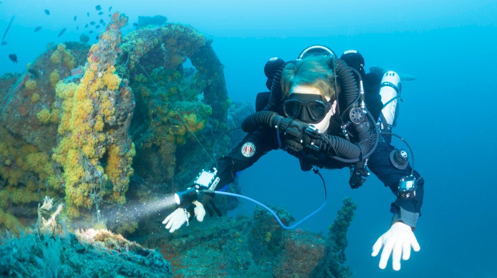 A scuba diver wearing a full wetsuit and diving mask explores an underwater shipwreck covered in colourful corals and marine growth, shining a torch on the wreckage.