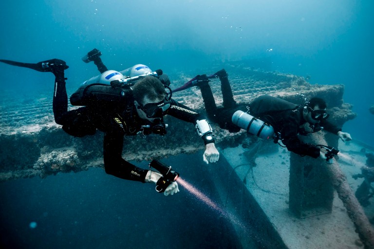 Two scuba divers in wetsuits and full kit explore an underwater shipwreck. They are using torches and gauges, surrounded by blue water and marine life on the metal structure.