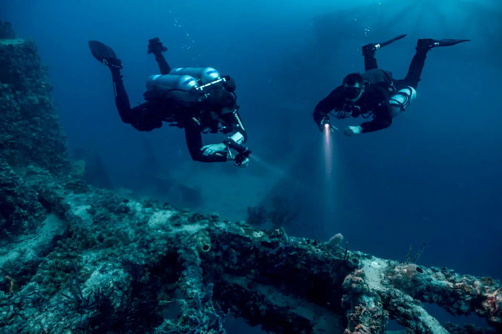 Two scuba divers with gear swim underwater near a coral-covered shipwreck, one holding a torch and pointing it at the wreck, surrounded by blue water and marine life.