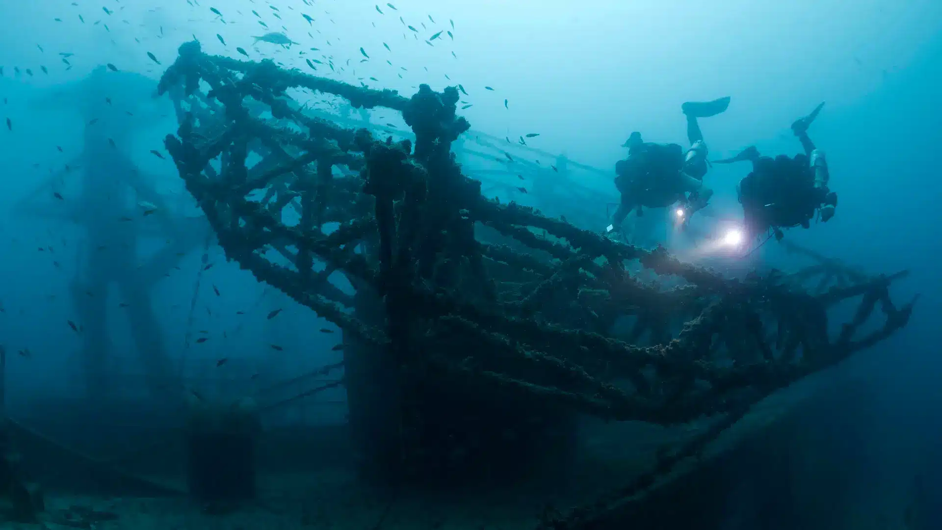 - N9BO℠ | Global Underwater Services Ltd Three scuba divers explore a large, rusted shipwreck underwater, surrounded by small fish and illuminated by the divers’ torches in the blue ocean depths.