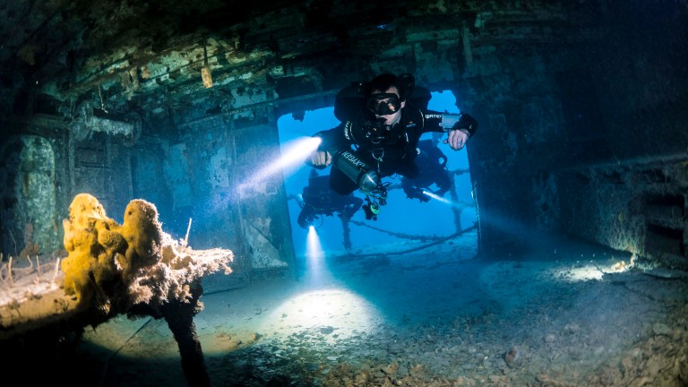 A scuba diver with a torch explores the interior of a sunken shipwreck underwater, surrounded by blue light and shadows, with debris and marine growth visible on the ship’s floor and walls.