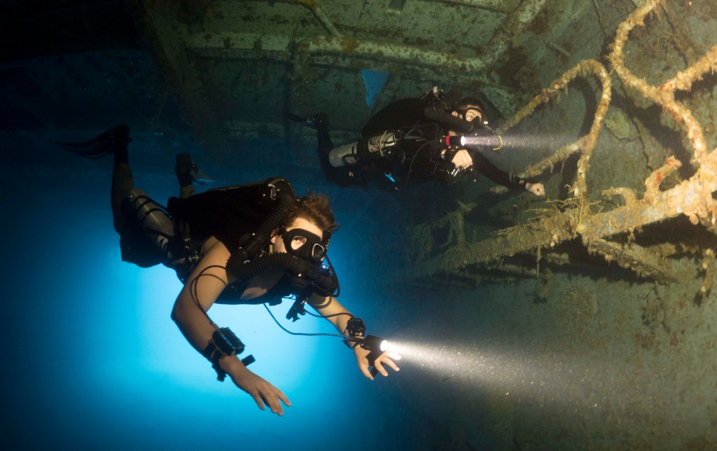 Two scuba divers with torches explore the dark interior of a sunken shipwreck underwater, surrounded by debris and illuminated by beams of light.