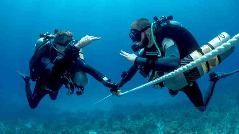 Two scuba divers underwater hold onto a rope, facing each other and using hand signals to communicate whilst surrounded by clear blue water and a seabed with some vegetation below.