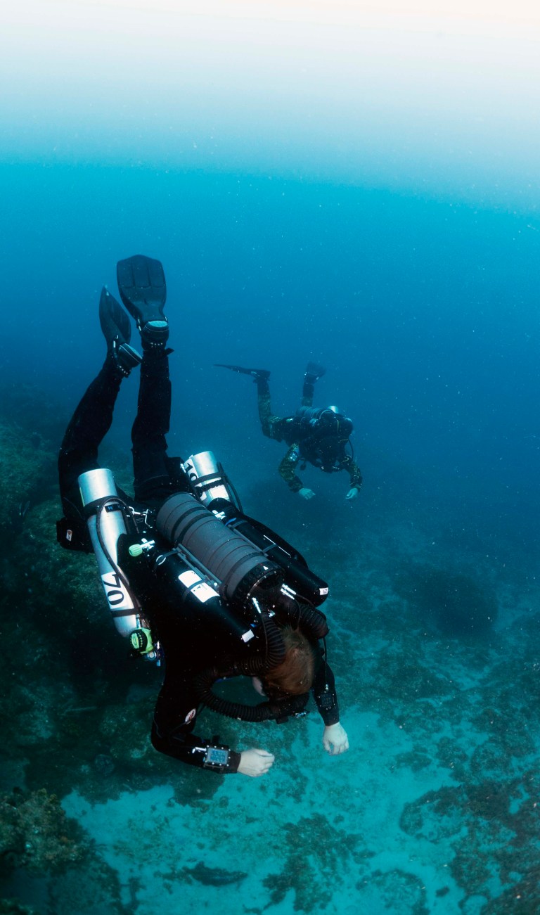 Two scuba divers in full kit swim underwater near a coral reef, surrounded by clear blue water. The divers appear to be exploring the sea, with one diver closer to the camera and the other further away.