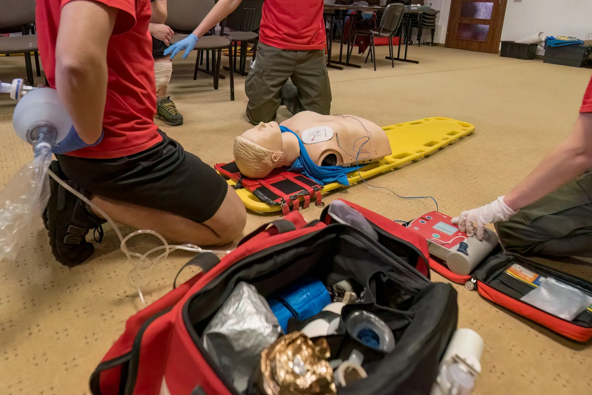Closeup of medical professionals performing CPR on a mannequin on the floor - N9BO℠ | Global Underwater Services Ltd Three people practise CPR and defibrillator use on a training manikin lying on a yellow stretcher, with medical supplies and equipment spread out in the foreground.