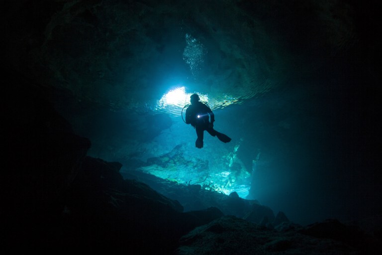 A scuba diver swims underwater inside a dark cave, illuminated by a beam of blue light coming from an opening above, with rocky surfaces visible in the cave interior.