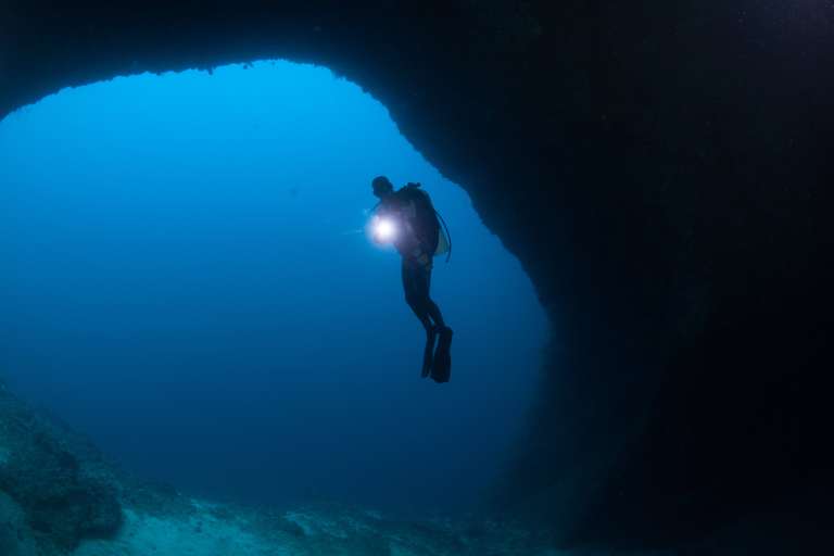 A scuba diver holding a torch swims near the entrance of a large underwater cave, surrounded by blue water and rocky formations.
