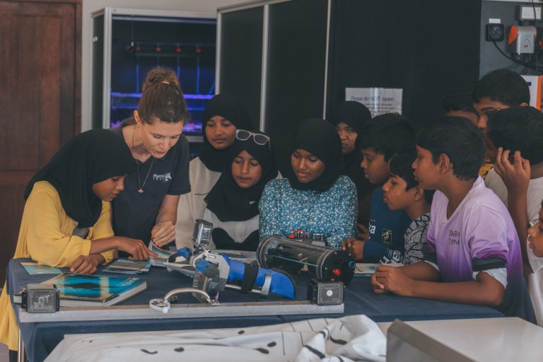 A group of students, some wearing hijabs, gather around a table with scientific equipment, watching a teacher demonstrate an experiment in a classroom or laboratory setting.