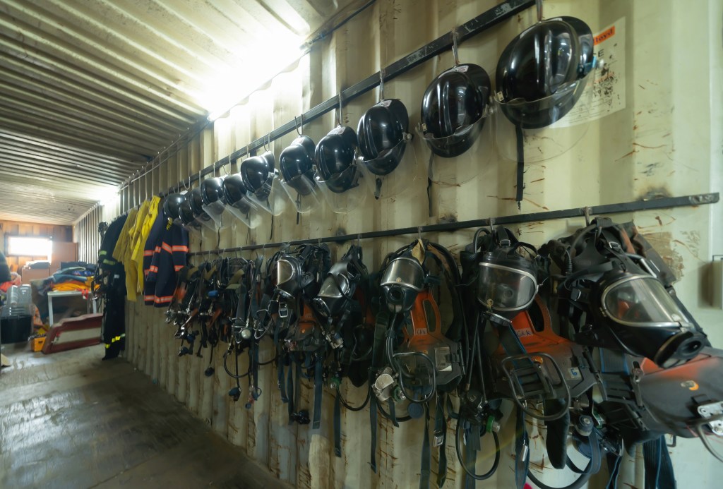 A row of black helmets and protective gear, including face masks and vests, hang neatly on hooks along the interior wall of a well-lit industrial container or storage area.