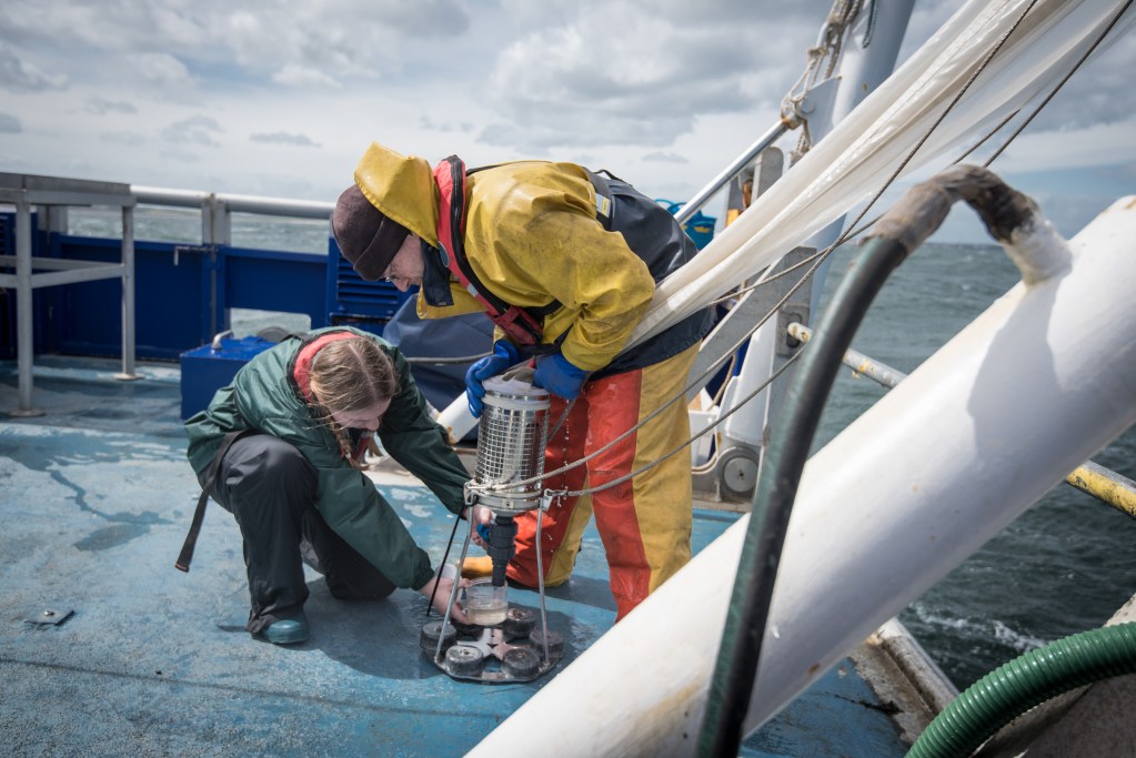Two people in weatherproof gear collect water samples with scientific equipment on the deck of a research boat at sea, working together under cloudy skies.