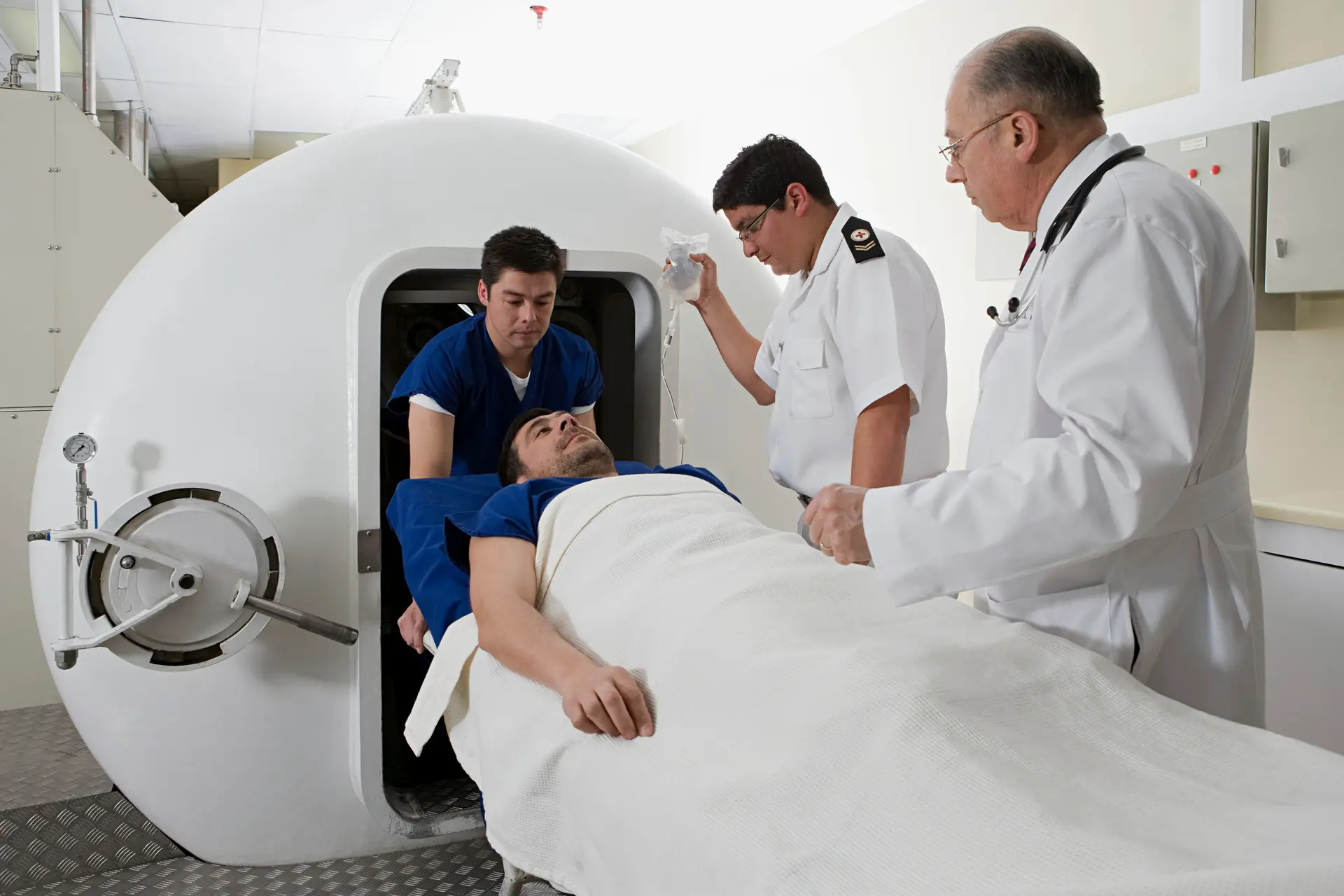A patient lies on a trolley being prepared to enter a hyperbaric oxygen therapy chamber, assisted by two medical staff and observed by a doctor.
