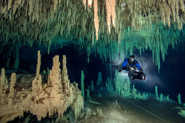 A scuba diver explores an underwater cave filled with stalactites hanging from the ceiling and stalagmites rising from the cave floor, illuminated by the diver’s torch.