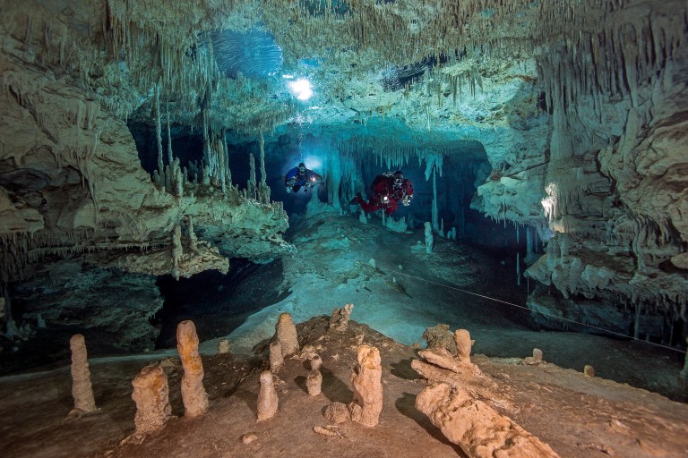 Two divers explore an underwater cave filled with stalactites hanging from the ceiling and stalagmites rising from the cave floor, illuminated by their torches and natural light from above.