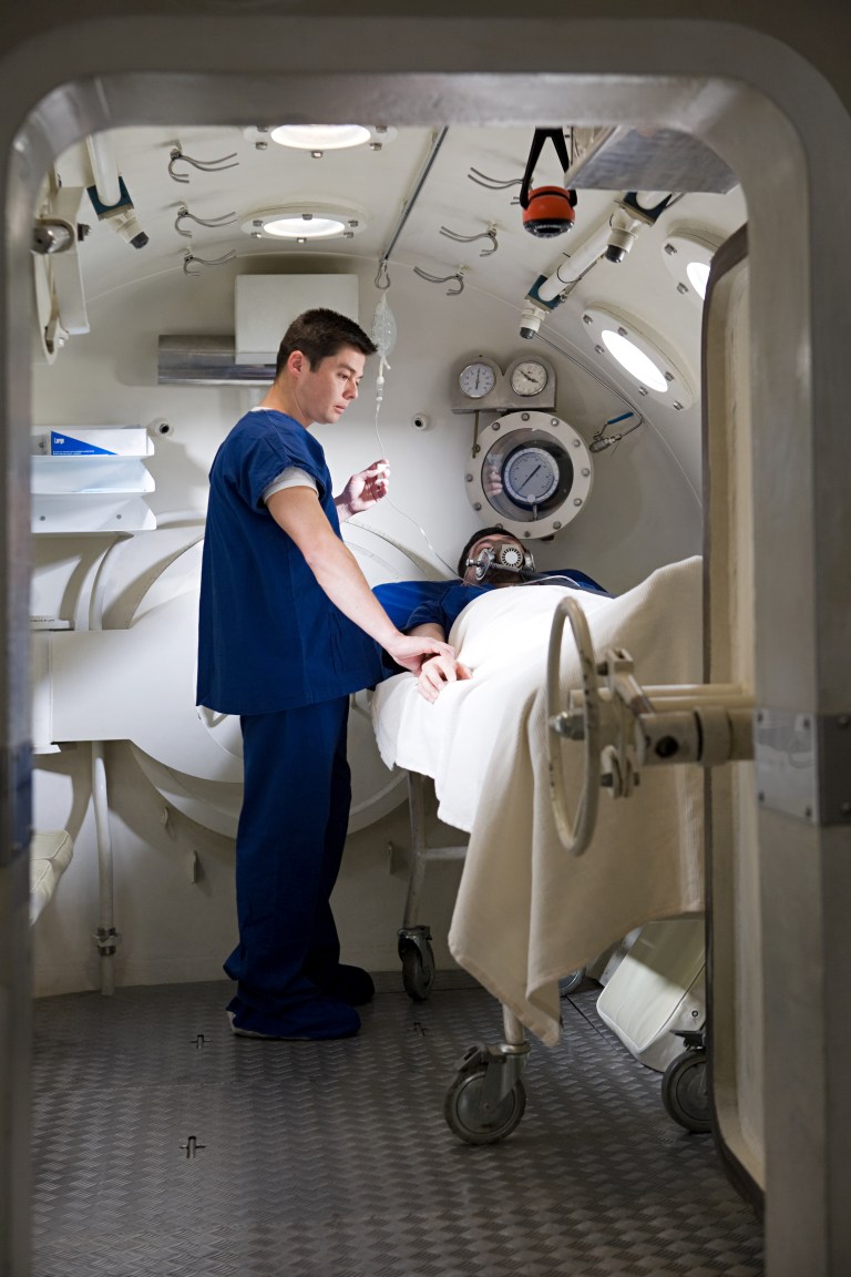 A healthcare professional in blue scrubs attends to a patient lying on a bed inside a hyperbaric oxygen chamber, with medical equipment and gauges visible on the chamber walls.