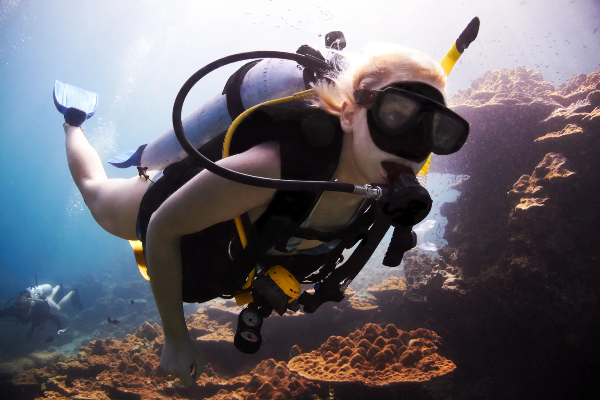 A person wearing scuba diving kit swims underwater near coral formations, surrounded by clear blue water and sunlight filtering from above.