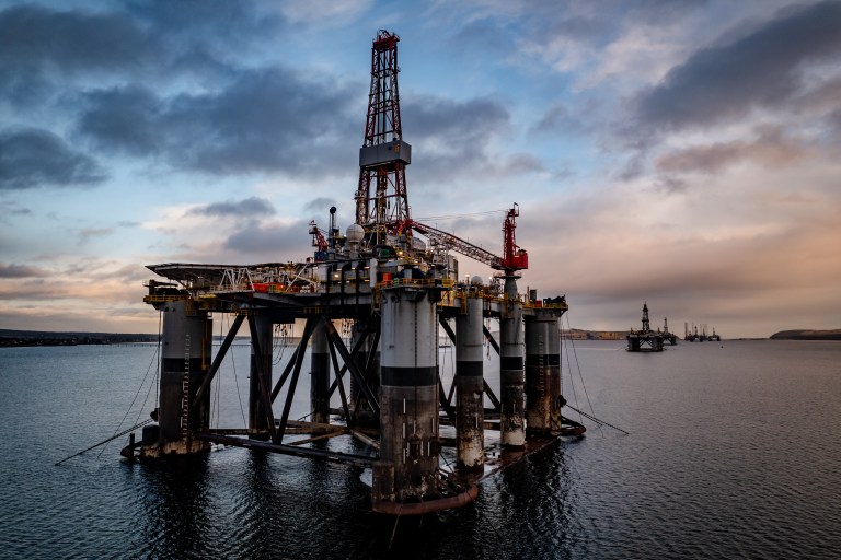 An offshore oil drilling platform stands in calm ocean waters under a cloudy sky at sunset, with another similar platform visible in the distance.