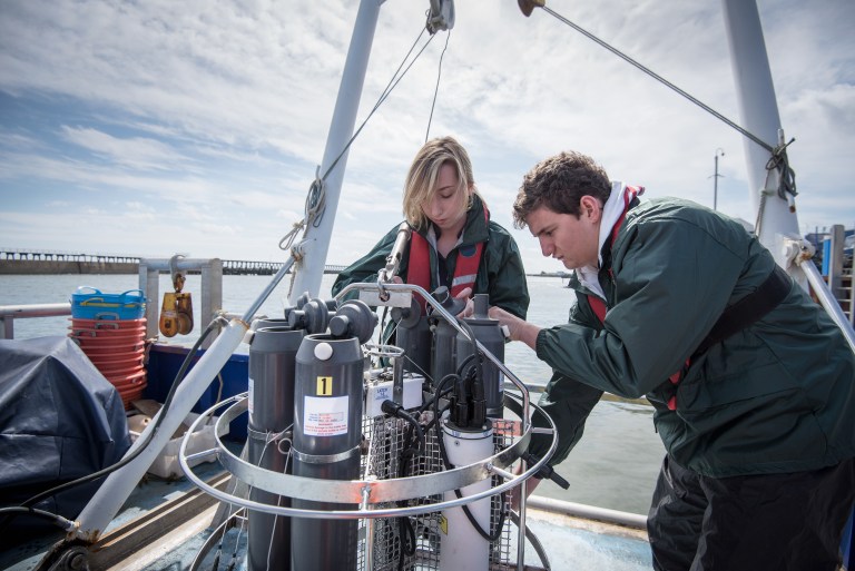 Two people in life jackets examine scientific equipment on a boat, likely preparing for or conducting marine research. The background shows calm water and a distant pier under a partly cloudy sky.