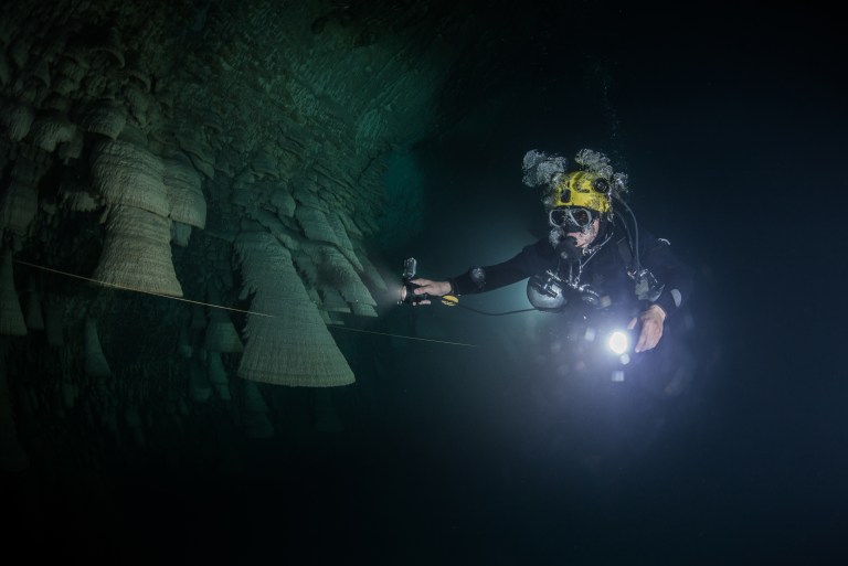 A scuba diver wearing a helmet with lights explores an underwater cave, holding a guideline and a torch. Large rock formations are visible on the cave wall. The scene is dark with the diver illuminated by their kit.