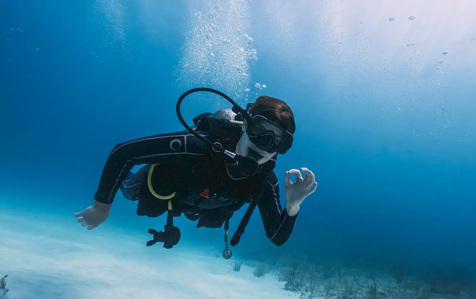 A scuba diver underwater wearing a black wetsuit and mask, making an OK hand signal whilst swimming above the sandy sea floor with some seagrass visible. Bubbles rise to the surface.
